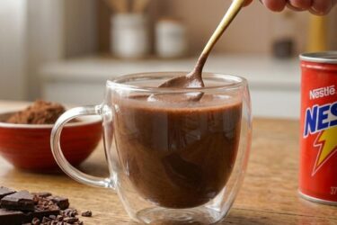 Caneca de porcelana com chocolate quente cremoso e denso, polvilhado com cacau em pó e canela em pau ao lado.