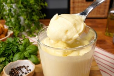 Pote de vidro com requeijão caseiro branco e cremoso, com uma faca passando em uma torrada ao lado.