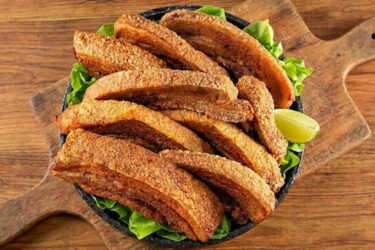 Pedaços de torresmo na cerveja bem dourados e com a pele pururucada e crocante, servidos em uma travessa com fatias de limão ao redor.