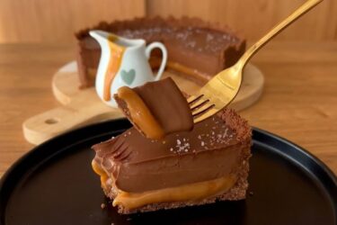 Fatia de torta de chocolate com caramelo deitada no prato, mostrando a base de biscoito, a camada espessa de caramelo puxa e o topo de ganache com flor de sal.