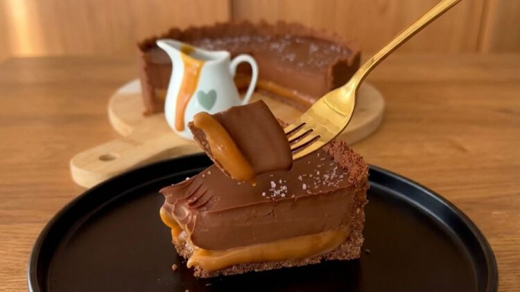 Fatia de torta de chocolate com caramelo deitada no prato, mostrando a base de biscoito, a camada espessa de caramelo puxa e o topo de ganache com flor de sal.