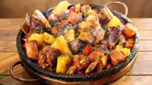 Panela de ferro servindo vaca atolada fumegante com pedaços de costela, mandioca macia, rodelas de linguiça e tomates-cereja por cima.
