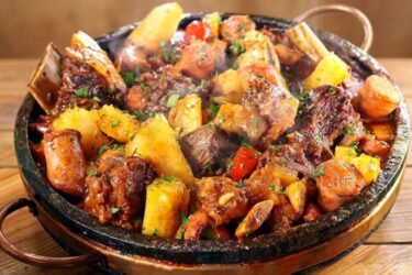 Panela de ferro servindo vaca atolada fumegante com pedaços de costela, mandioca macia, rodelas de linguiça e tomates-cereja por cima.