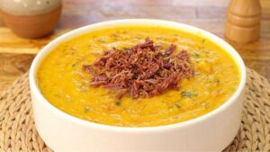 Tigela de cerâmica com caldo de abóbora com carne seca cremoso bem alaranjado, com carne seca desfiada e cheiro-verde por cima.