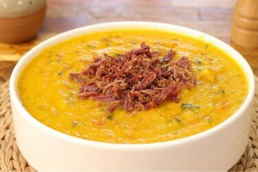 Tigela de cerâmica com caldo de abóbora com carne seca cremoso bem alaranjado, com carne seca desfiada e cheiro-verde por cima.