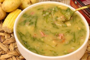 Tigela de cerâmica com caldo verde cremoso, mostrando fatias de calabresa, pedacinhos de bacon e tiras de couve, com um pãozinho ao lado.