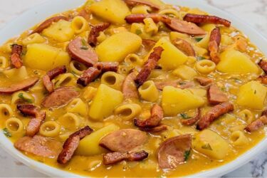 Prato fundo com sopa de feijão bem cremosa e grossa, com rodelas de calabresa, cubos de batata, macarrão e cheiro-verde, salpicada com bacon crocante.