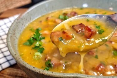 Tigela cheia de caldo de mandioca com bacon e calabresa fumegante, decorada com cheiro-verde picadinho por cima.