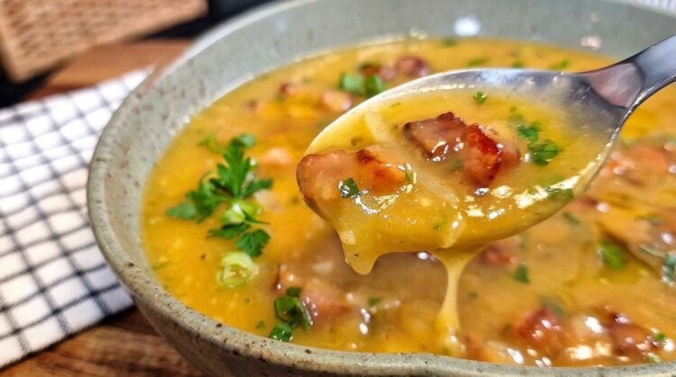 Tigela cheia de caldo de mandioca com bacon e calabresa fumegante, decorada com cheiro-verde picadinho por cima.