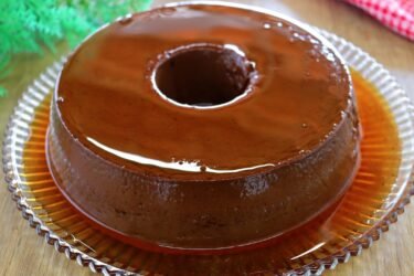 Pudim de chocolate sem ovo desenformado em um prato, coberto com calda de caramelo brilhante e fatia cortada ao lado.