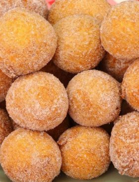 Cesta de vime cheia de bolinho de chuva com massa de banana, polvilhados com açúcar e canela, ao lado de uma xícara de café