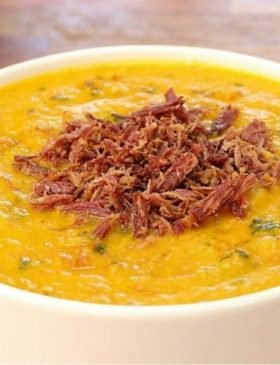Tigela de cerâmica com caldo de abóbora com carne seca cremoso bem alaranjado, com carne seca desfiada e cheiro-verde por cima.
