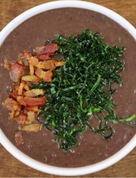 Caneca de cerâmica cheia de caldo de feijão preto bem cremoso, finalizado com couve crispy crocante e cubos de bacon frito por cima.