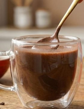 Caneca de porcelana com chocolate quente cremoso e denso, polvilhado com cacau em pó e canela em pau ao lado.