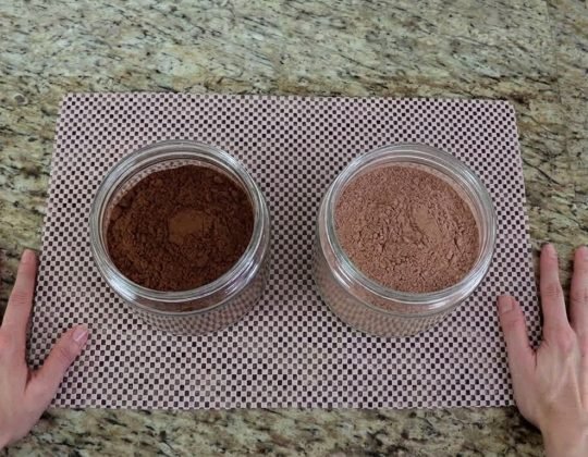 Chocolate em pó x achocolatado qual usar nas receitas Dois potes na bancada da cozinha, um com chocolate em pó escuro e outro com achocolatado claro