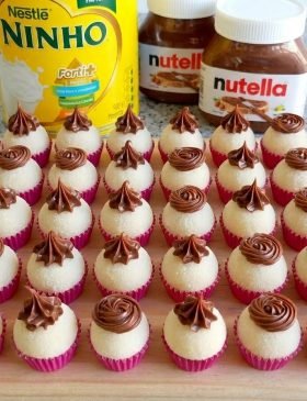 Brigadeiros de leite Ninho dispostos em forminhas, cobertos com leite em pó e decorados com rosetas de Nutella no topo.