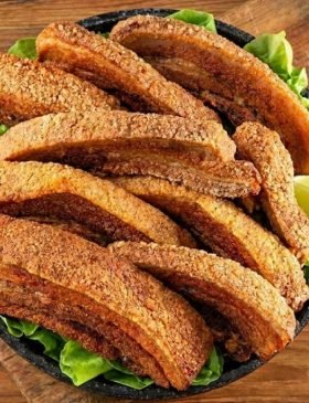 Pedaços de torresmo na cerveja bem dourados e com a pele pururucada e crocante, servidos em uma travessa com fatias de limão ao redor.