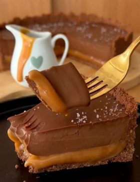 Fatia de torta de chocolate com caramelo deitada no prato, mostrando a base de biscoito, a camada espessa de caramelo puxa e o topo de ganache com flor de sal.