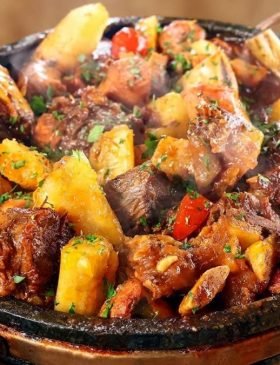 Panela de ferro servindo vaca atolada fumegante com pedaços de costela, mandioca macia, rodelas de linguiça e tomates-cereja por cima.